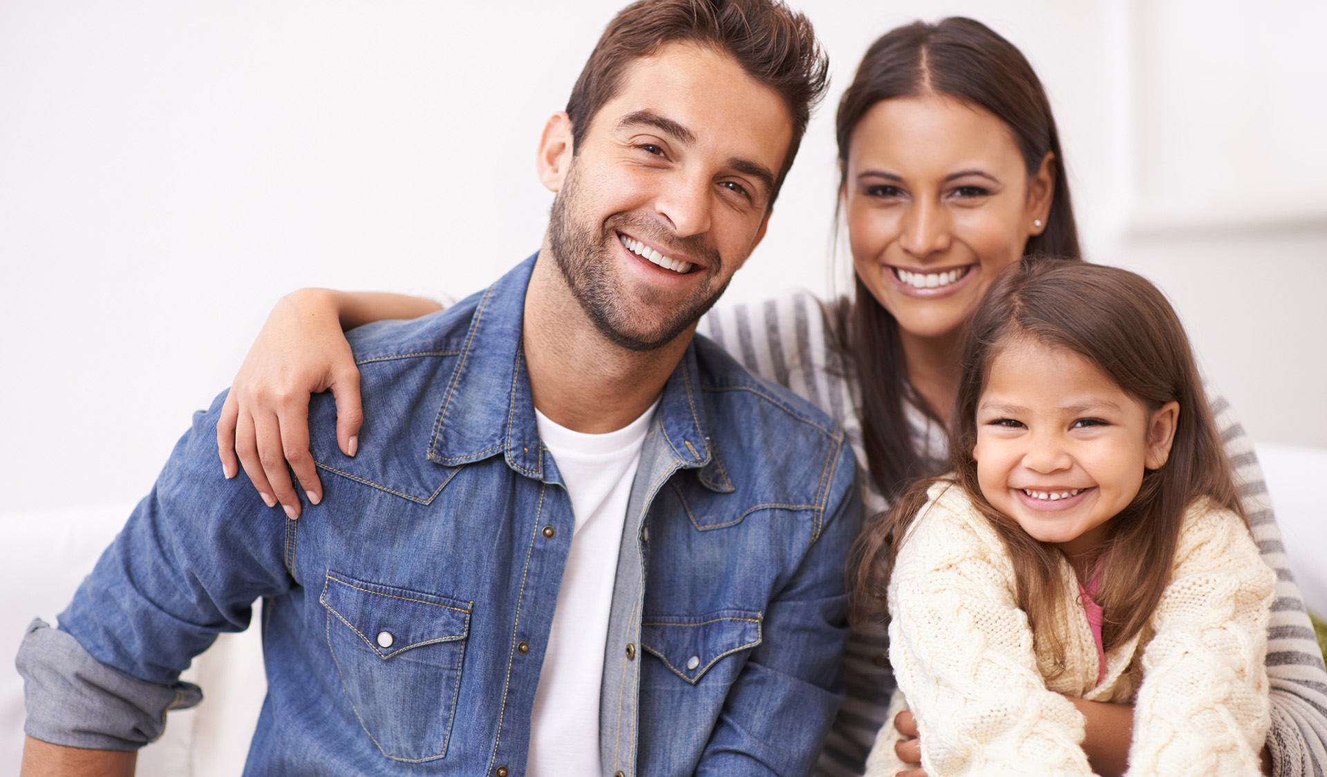 Image of family of three smiling showing off their teeth. Man positioned on left and woman holding female toddler on right.